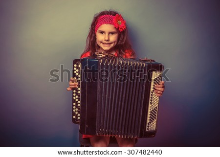 a girl of seven European appearance brunette in a bright dress plays accordion on gray background, music, talent retro