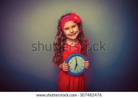 a girl of seven European appearance brunette in a bright dress holding a blue watch on gray background, time, smile retro
