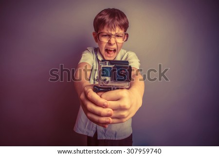 A boy of about ten European appearance in light brown shirt and glasses holding a small camera action and shouts against the gray background, video  GoPro, screaming, go pro camera retro