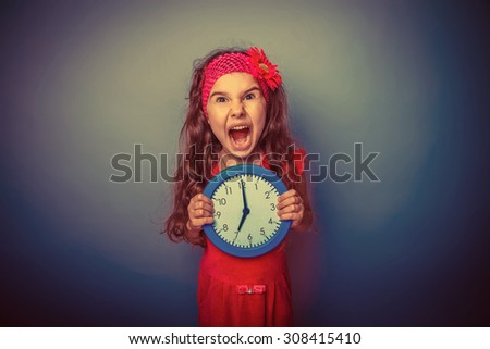 a girl of seven European appearance brunette in a bright dress holding a blue clock and shouting his mouth open on a gray background, the time, the lack of retro