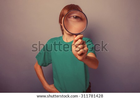 European-looking boy of ten years is looking through a magnifying glass, a keen eye on gray background retro