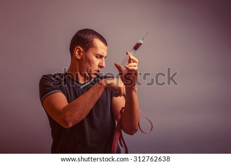 male of European appearance tightens the harness on one hand holds the syringe in the other hand, a drug addict on a gray background retro