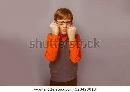 European-looking boy of ten  years  in glasses frowning, unhappy shows fists on a blue background retro