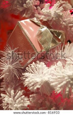 A snow covered pink gift box with a silver ribbon sits on the branch of a flocked Christmas tree with pink lights.