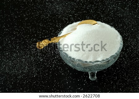 Pure snowy white sugar in a vintage crystal bowl with a gold spoon sitting on a sugar covered black textured background.