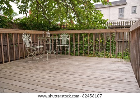 A weathered wood deck with an old iron bistro table and chairs placed in the corner, nestled in the shade of a tree in the back yard.
