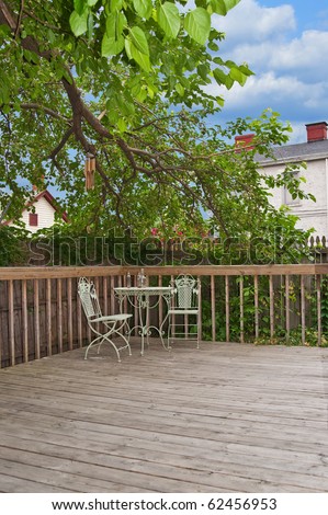 A weathered wood deck with an old iron bistro table and chairs placed in the corner, nestled in the shade of a tree in the back yard.