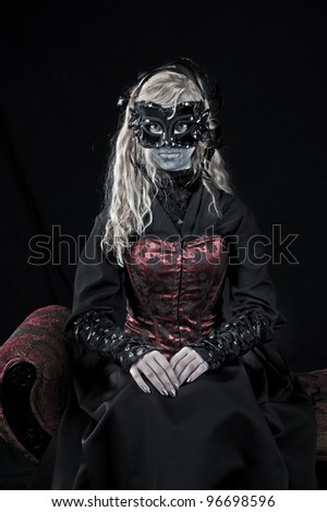 Goth style blonde vampire girl is wearing vintage style dark clothing and a mask while posing in a seated position.
