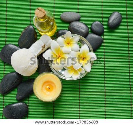 frangipani in bowl with massage oil and black stones on green bamboo mat