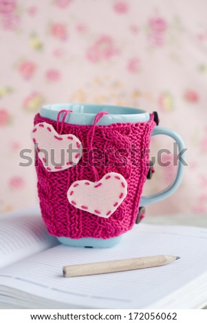 Blue cup in pink sweater with felt hearts standing on an open notebook