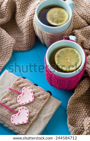 Two blue cup of tea in knitted sweater with hearts felt standing next to the old notebook and blanket knitted on blue background