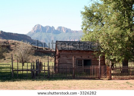Old cabin in the shade of a large tree with mountains in the background.