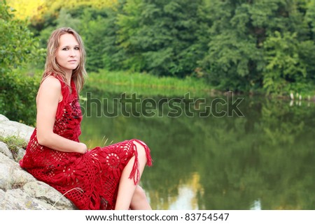 Beautiful woman wrapped in a red shawl sits on the stones by the coast river