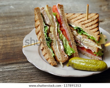 Photo of a club sandwich made with turkey, bacon, ham, tomato, cheese, lettuce, and garnished with a pickle.