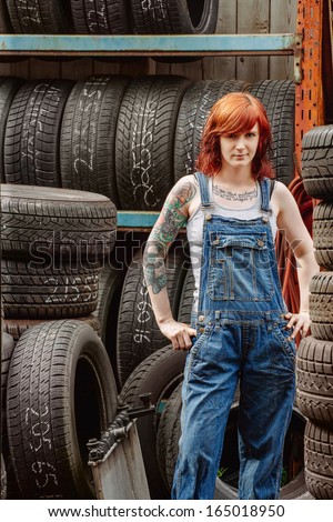 Photo of a young beautiful redhead mechanic wearing overalls and standing in an old garage. Attached property release is for arm tattoos.