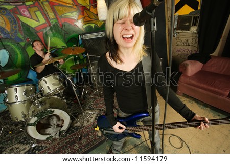 Female bass player screaming into a microphone.  Focus on singers face - drummer blurry in the background.  Shot with slow shutter speed and strobes - motion blur visible in some areas.