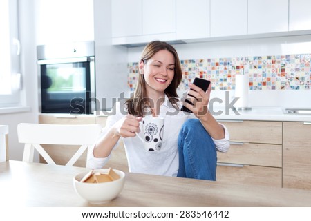 Young beautiful woman using cell phone and having a coffee in the kitchen