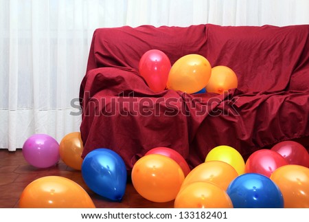 Many colorful party balloons laying in the floor and on a red couch in a living-room
