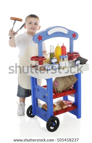 A young preschooler by his vendor stand holding a hot dog on the end of his tongs.  The stand\'s signs left blank for your text.  On a white background.