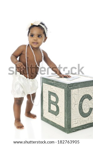 A beautiful baby girl standing with support.  She wears a white hair bow, long strands of beads and silky bloomers.  On a white background.