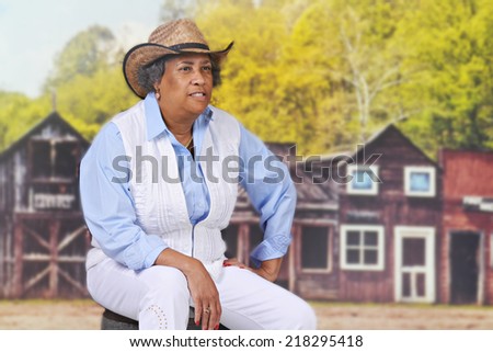 A senior adult cowgirl sitting on an old barrel near an old western town.