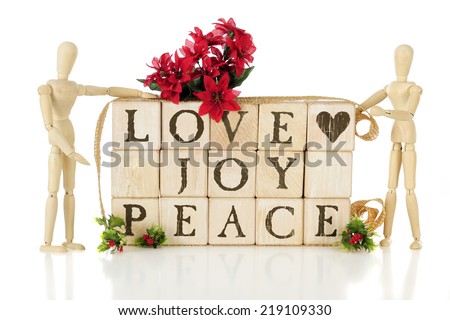 Rustic alphabet blocks arranged to say LOVE, JOY, PEACE.  They are flanked by two wooden mannequins and adorned with Christmas holly and poinsettias.  On a white background.