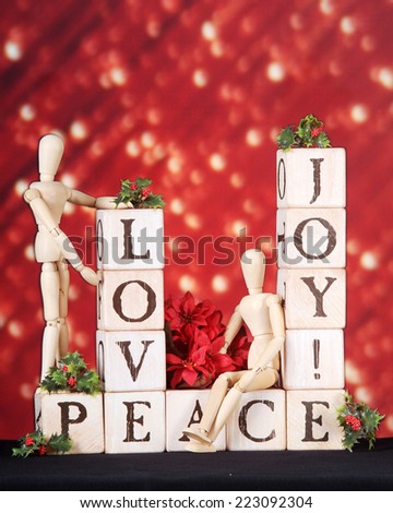 Love, joy and Peace spelled out in rustic Alphabet blocks.  They\'re adorned by two mannequins, holly and a small bouquet of red poinsettias on a red background of festive lights.
