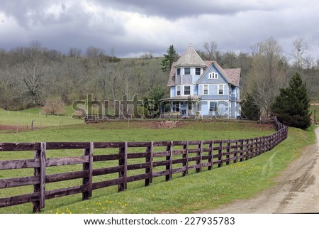 A rustic fence and dirt road by an old Victorian home under an overcast sky in the early spring.