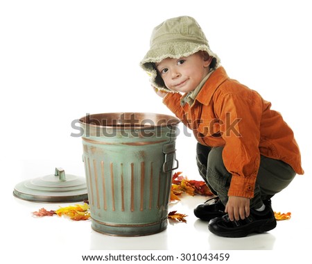 An adorable preschooler in fall colors squatting beside a small trash can as he cleans up fallen autumn leaves.  On a white background.