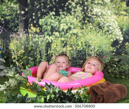 Two preschool sisters looking at the viewer as they relax outside in their filled and inflated kiddie pool.