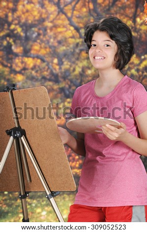 A pretty young teen looking up at the viewer as she paints on an easel outside in the fall.