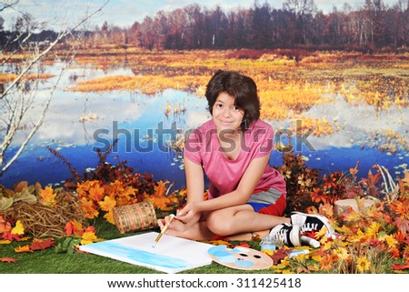 A pretty young teen in the fall  looking up at the viewer as she begins to paint the scene around her as she sits at the edge of pond.