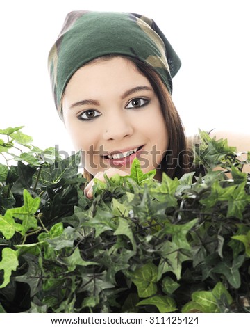 Close-up of a beautiful teen girl in a camouflage head scarf peeking over thick, green ivy.  On a white background.