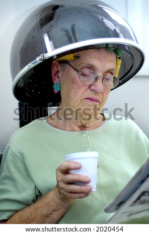 Senior woman under beauty parlor hair dryer, reading paper and drinking coffee.