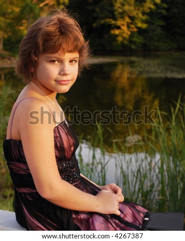 A happy young girl in a ballroom gown sits by a pond at sunset looking toward the viewer.
