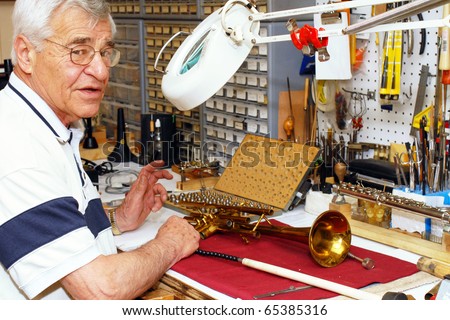 A senior man at work in his musical instrument repair shop where he\'s working on a trumpet.