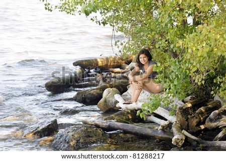 A pretty young teen tucked under summer foliage by the rocks and fallen logs of a rough lakeshore.