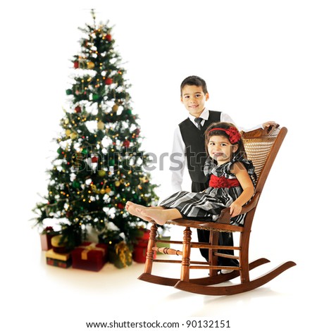 Dressed-up siblings posing near a Christmas tree in an old rocking chair.  On a white background.