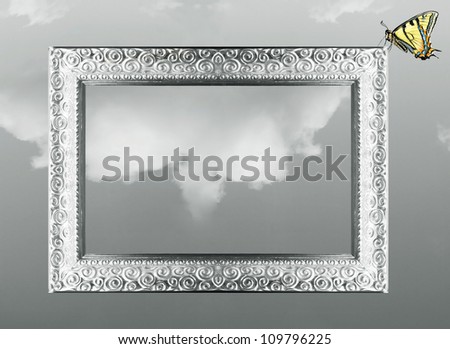 Imaginative fantasy background of a silver baroque frame with a beautiful colored butterfly on the angle of it on black and white cloudy sky on the background