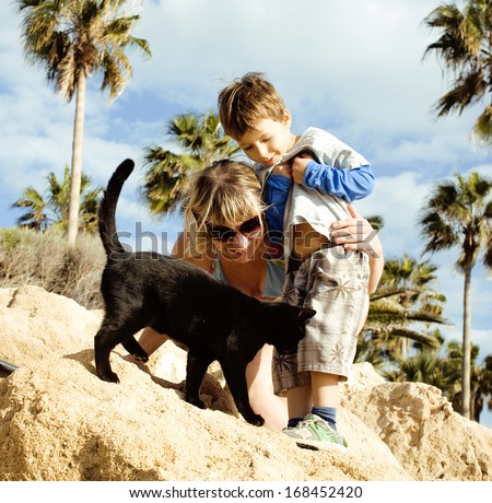 grandmother with grandson on cyprus beach among palms and rock playing with black cat