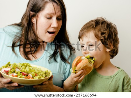 fat woman holding salad and little cute boy with hamburger teasing, real family