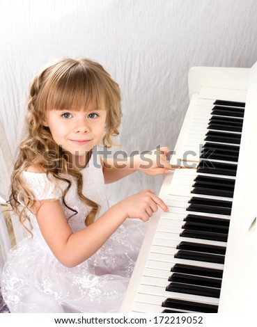 little beautiful girl with blond long curls in white fashion dress playing on white piano