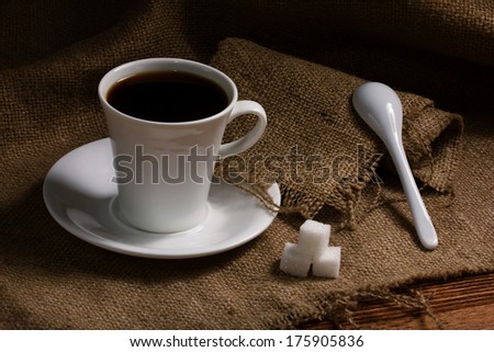 white ceramic cup with espresso coffee on saucer, ceramic spoon and cubes sugar on burlap on wooden table