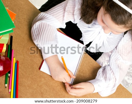 beauty little girl  in school form writing in copybook, near -  red apple, many colorful books and pencils, top view