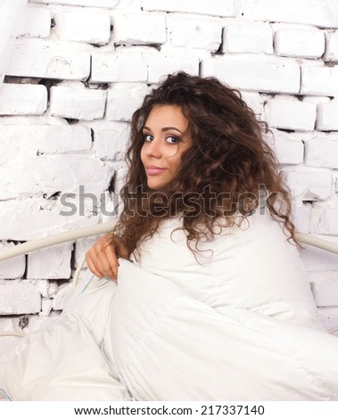beautiful  young woman with long dishevelled curls sitting  under duvet on white bricky wall background