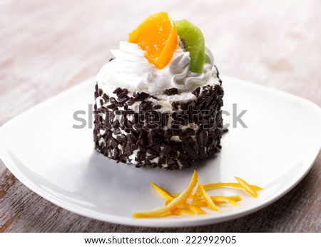 cake with cream, chocolate crumb, slices of tropical fruits and orange skin  on white plate on wooden surface
