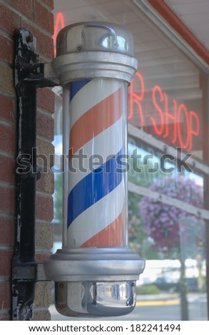 An old-style barber pole on the front of a small town barber shop, with the neon sign and a reflection of the town street in the window