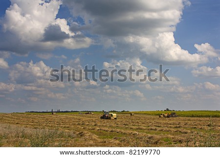Farming on a large horizon featuring a hay bailer and hay lifter under a big cloudy sky