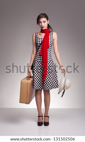 young lady with retro strap plaid dress carrying a suitcase on her hand and a hat on her other hand,waiting for her vehicle on grey background
