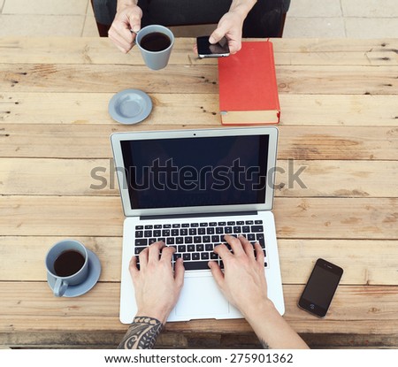 young guy with tattooed arm using a laptop while his friend using a smartphone